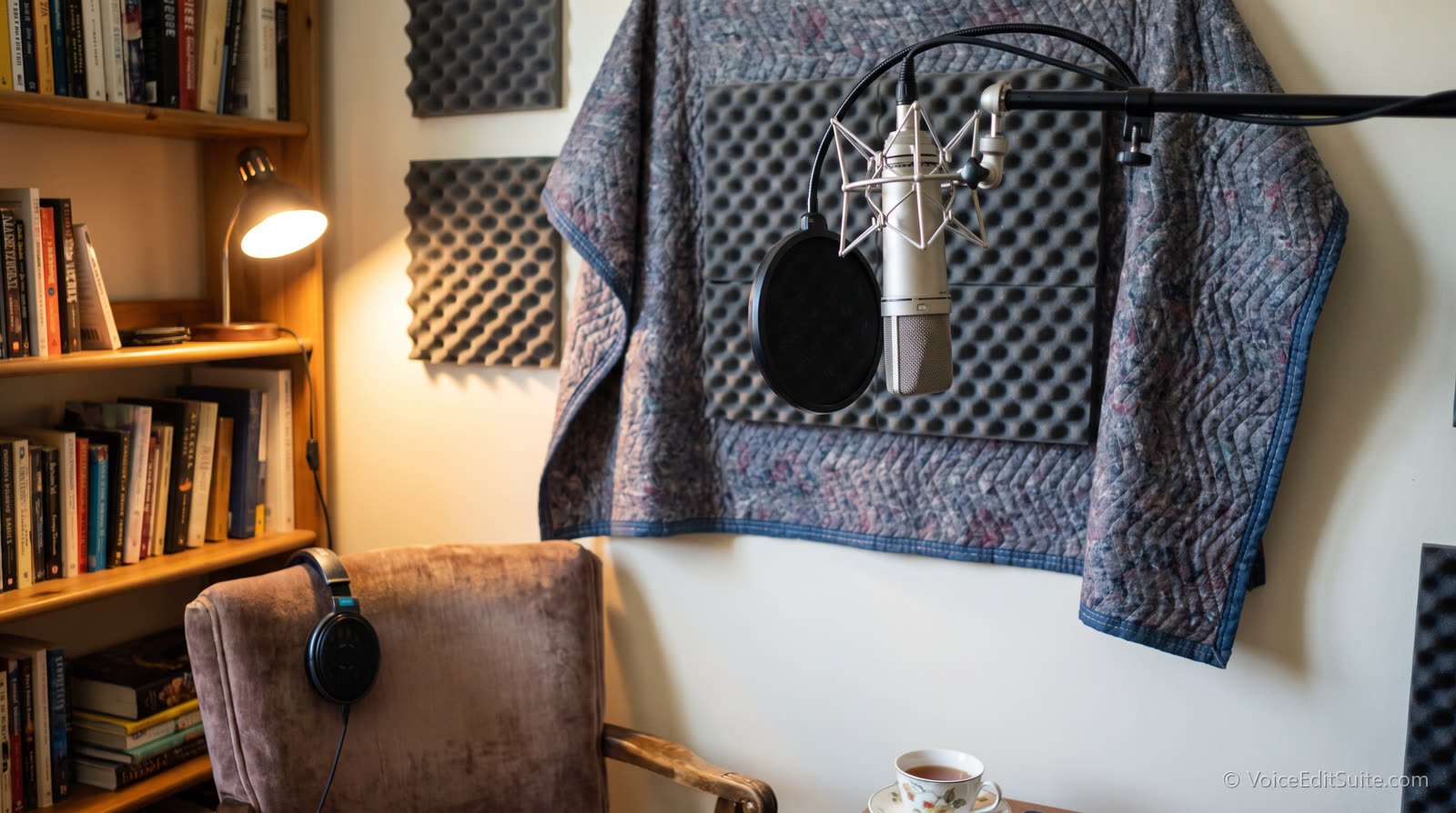 A budget home voice over corner: a condenser microphone on a boom arm in front of a moving blanket and acoustic foam, with a bookshelf and armchair nearby